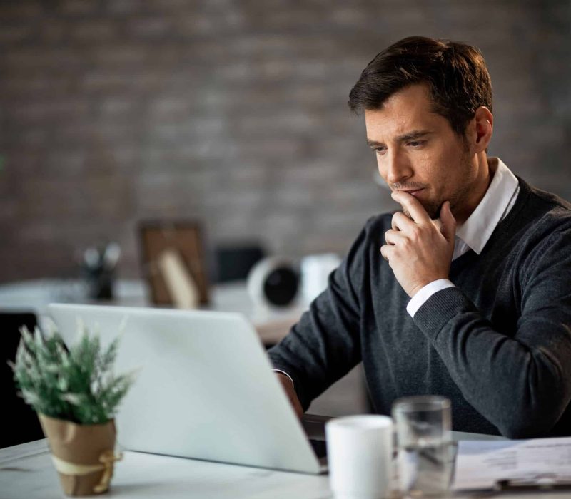 Smiling businessman using laptop and contemplating while working at his desk in the office.